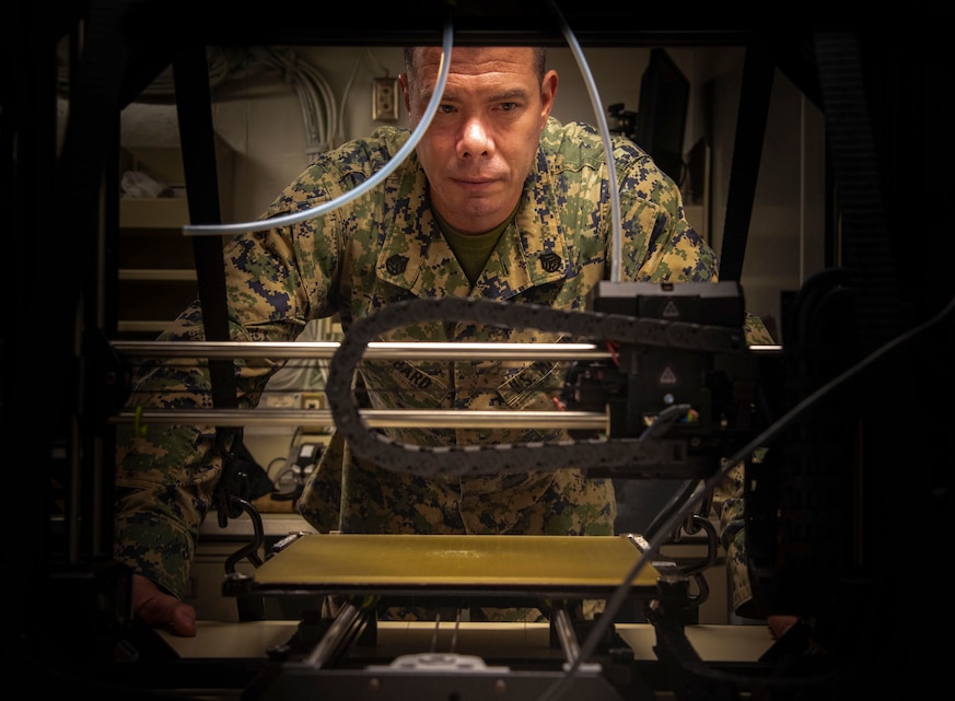 A U.S. Marine observes a 3D printer as it prints an equipment part used aboard USS New Orleans (LPD 18), Aug. 24.